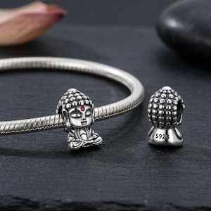 Puede incluir: Una pulsera de cadena de serpiente de plata con un dije de Buda que tiene una pequeña piedra roja en la frente. Un segundo dije de Buda idéntico se encuentra al lado, mostrando el sello S925 grabado en la base de la figura de plata.