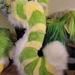 May include: A plush tail in a spiral design. The tail has alternating sections of bright green and pale yellow, with white fur at the ends. The tail is part of a costume or cosplay item.
