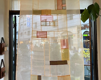 May include: A sheer curtain with a patchwork design. The curtain is rectangular and features a grid of beige, brown, pink, and green patches. It hangs in front of a window, allowing a view of the outside through the sheer fabric.