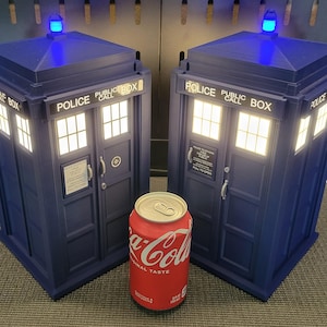 Könnte beinhalten: Zwei marineblaue Police Box-Repliken mit beleuchteten Fenstern und blauen Oberlichtern. Die Repliken tragen die Aufschrift "POLICE PUBLIC CALL BOX". Eine rote Coca-Cola-Dose steht vor den Repliken.