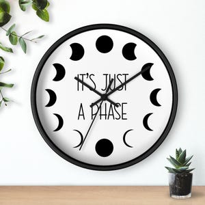 May include: Black and white wall clock with a black frame and a white face. The clock has a moon phase design and the text "It's just a phase".