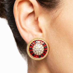 May include: A gold-toned, round earring with a red enamel center and a rope-like border. The center features a cluster of clear crystals. Black text on the red enamel reads "NOSTIBUTTER". The earring is worn on the ear.