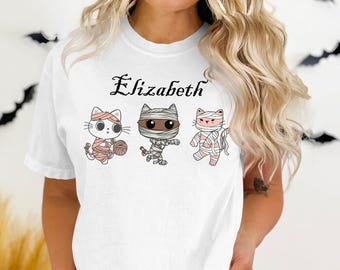 Pode incluir: Camiseta branca com três gatos de desenho animado vestidos com fantasias de Halloween. Os gatos são retratados como uma múmia, um gato com bandagens e um gato com novelo de lã. O nome "Elizabeth" está escrito acima dos gatos em uma fonte decorativa.