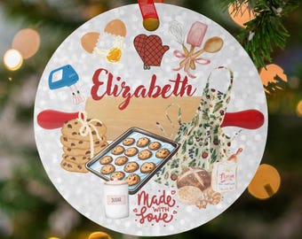 May include: Circular Christmas decoration with a baking theme. It features illustrations of cookies, an apron, and baking tools. The name "Elizabeth" is written in red script, and the phrase "Made with Love" is also included.