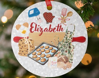 May include: A circular Christmas ornament with a red ribbon. It features baking-related illustrations such as a mixer, oven mitt, rolling pin, and cookies. The name "Elizabeth" is written in red script across the ornament.