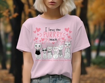 Peut inclure: T-shirt rose clair avec l'inscription "I love you purrry much" et des illustrations de chats. Les chats sont vêtus de différentes tenues, dont un smoking, des lunettes de soleil et des écharpes. Les noms "Mother", "Danny" et "Mommy" sont imprimés en dessous.