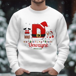 Könnte beinhalten: Weißes Sweatshirt mit Rundhalsausschnitt und weihnachtlichem Design. Das Design zeigt einen großen roten Buchstaben "D" mit Weihnachtsmütze, einen schwarzen Gürtel mit goldener Schnalle und den Namen "Dewayne" in roter Schrift. Ein Weihnachtsmann und ein Rentier sind ebenfalls abgebildet.