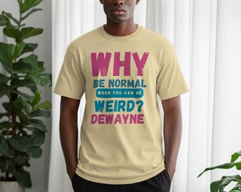 Puede incluir: Una camiseta de color beige claro con el texto "WHY BE NORMAL WHEN YOU CAN BE WEIRD? DEWAYNE" en rosa, azul y turquesa. La camiseta es de manga corta y la lleva una persona.