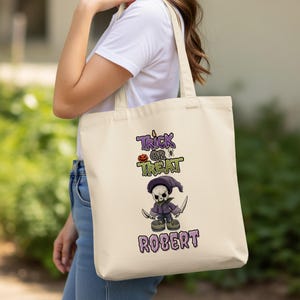 Puede incluir: Bolso tote beige con un diseño de Halloween. El diseño incluye las palabras "Trick or Treat" sobre un personaje de esqueleto de dibujos animados con ropa morada, sosteniendo cuchillos, con el nombre "Robert" debajo.