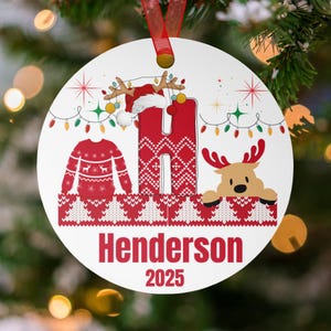 May include: A white Christmas ornament with a red ribbon. The ornament features a red letter "H" with a Santa hat and reindeer antlers, a red sweater, and a reindeer peeking over a patterned border. The name "Henderson" and the year "2025" are printed in red.