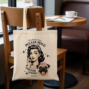 May include: A cream-coloured tote bag with a vintage illustration. It features a woman with a retro hairstyle and a pug wearing a pink bow. Text on the bag reads "Elizabeth Anne, PLEASE HOLD, SARCASM LOADING, Baxter."