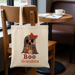May include: A cream tote bag with a patchwork ghost design and an orange bow. The ghost has a black face and is made of brown, orange, and grey fabric squares. The words "Boo Grandma" are printed in orange below the ghost, perfect for Halloween.