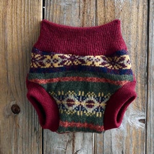 May include: A knitted dog diaper with a fair isle pattern. The diaper features a burgundy waistband and leg openings, with a green, blue, yellow, and burgundy patterned body. The garment is displayed against a rustic wooden background.