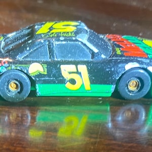 Puede incluir: Un pequeño coche de carreras de juguete con carrocería negra, detalles verdes y el número "51" en amarillo. El coche tiene texto "Mello Yello" y "Eagle Fuel". Las ruedas son negras con centros dorados y el coche tiene un alerón.