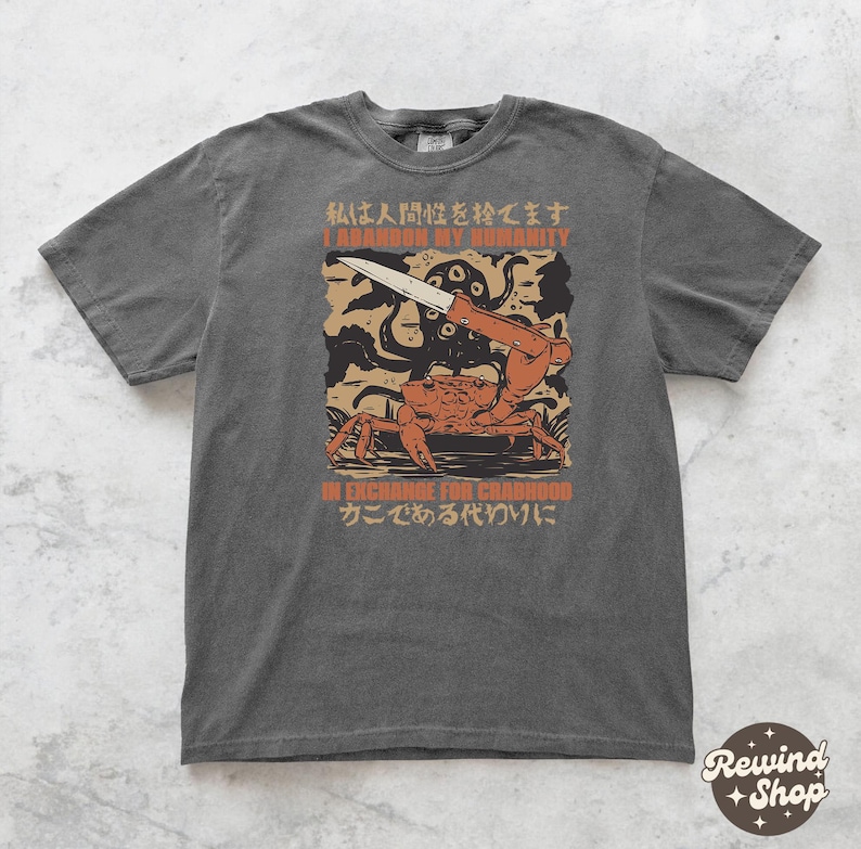 May include: Dark grey t-shirt featuring a graphic of a crab holding a knife, with an octopus and text that reads "I Abandon My Humanity In Exchange For Crabhood". The shirt has a vintage, distressed look.