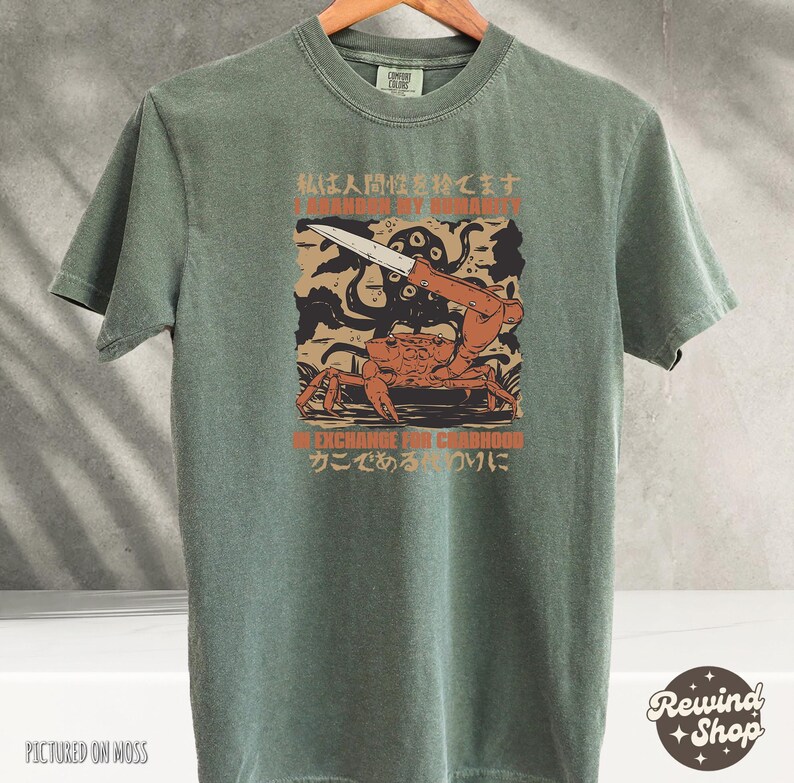 May include: Olive green t-shirt with a graphic featuring a crab holding a knife, an octopus, and Japanese text. The text reads "I abandon my humanity" and "In exchange for crabhood". The shirt is hanging on a wooden hanger.