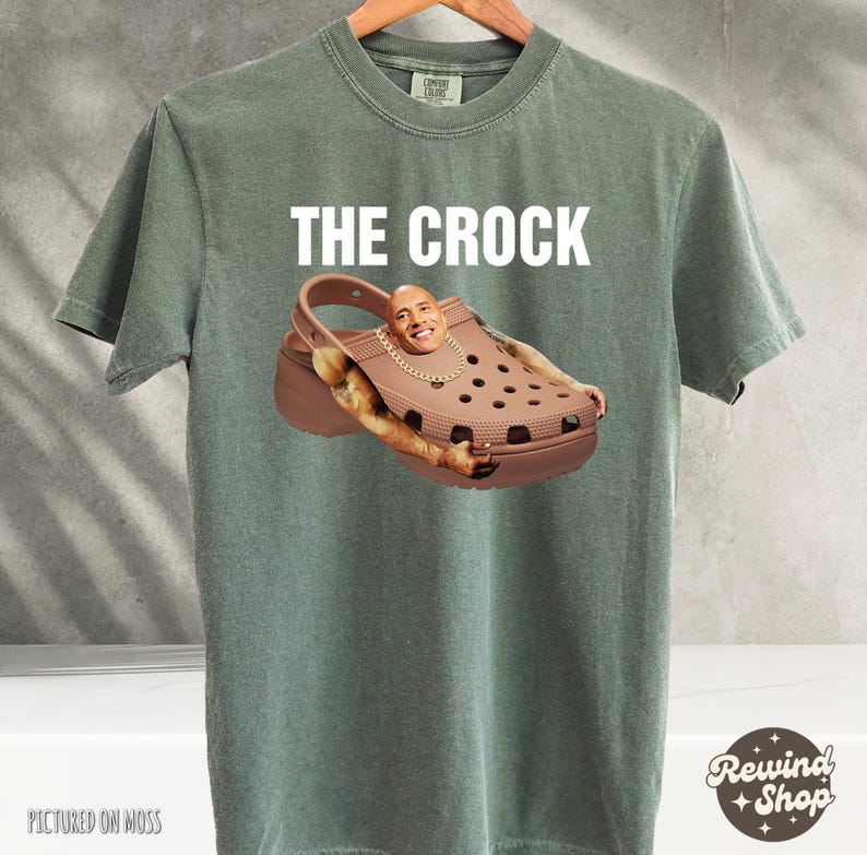 May include: A green t-shirt with the text "THE CROCK" printed on the front. The image shows a brown and black Croc shoe with a man's face inside the shoe.