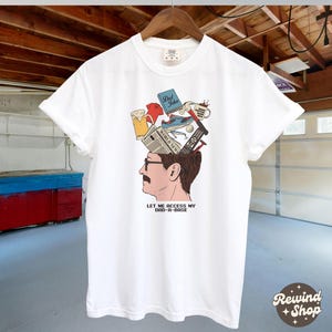May include: White t-shirt featuring a graphic of a man's head with various items emerging from it, including a beer, a shoe, and a remote control. The text "LET ME ACCESS MY DAD-A-BASE" is printed below the graphic.