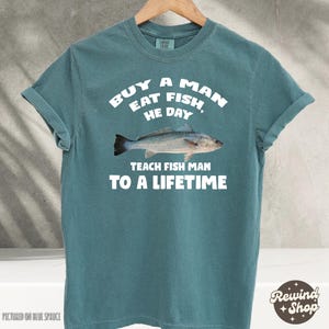 May include: A teal blue t-shirt with a white graphic print of a fish and the text "Buy a man eat fish, he day teach fish man to a lifetime".