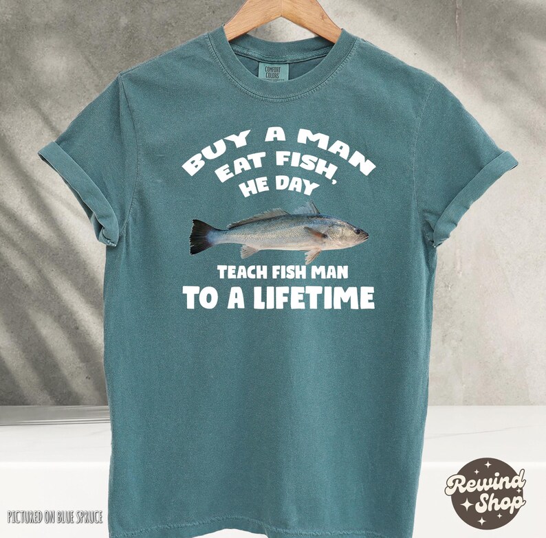 May include: A teal blue t-shirt with a white graphic print of a fish and the text "Buy a man eat fish, he day teach fish man to a lifetime".