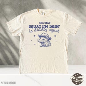 May include: A white t-shirt with a blue graphic of a possum wearing a cowboy hat. The text "THE ONLY SQUAT I'M DOIN' is diddly squat" is printed on the shirt.