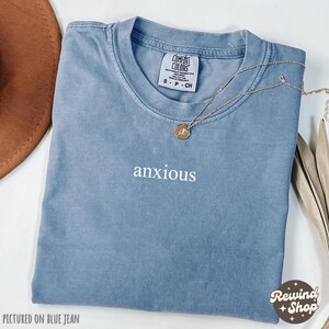 May include: A blue t-shirt with the word "anxious" embroidered on the front. The shirt is pictured on a blue jean background.