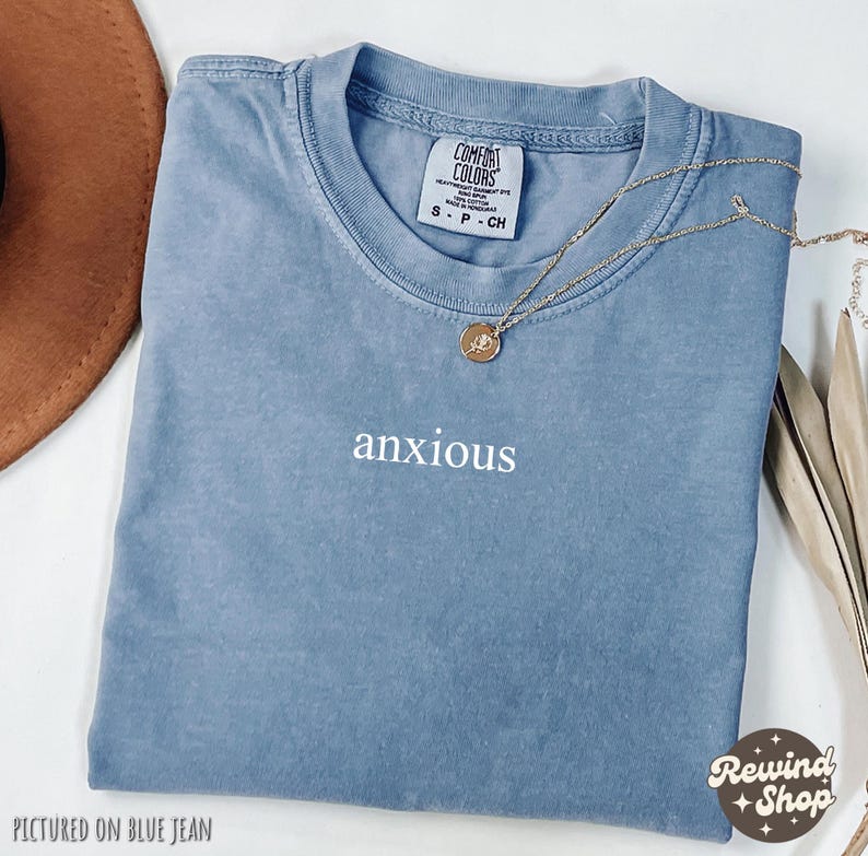 May include: A blue t-shirt with the word "anxious" embroidered on the front. The shirt is pictured on a blue jean background.