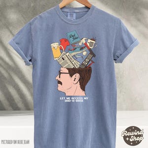 May include: Blue t-shirt with a graphic of a man's head, open to reveal various items like a beer, a remote, and a mug. The text "LET ME ACCESS MY DAD-A-BASE" is below the image. The shirt is displayed on a hanger.