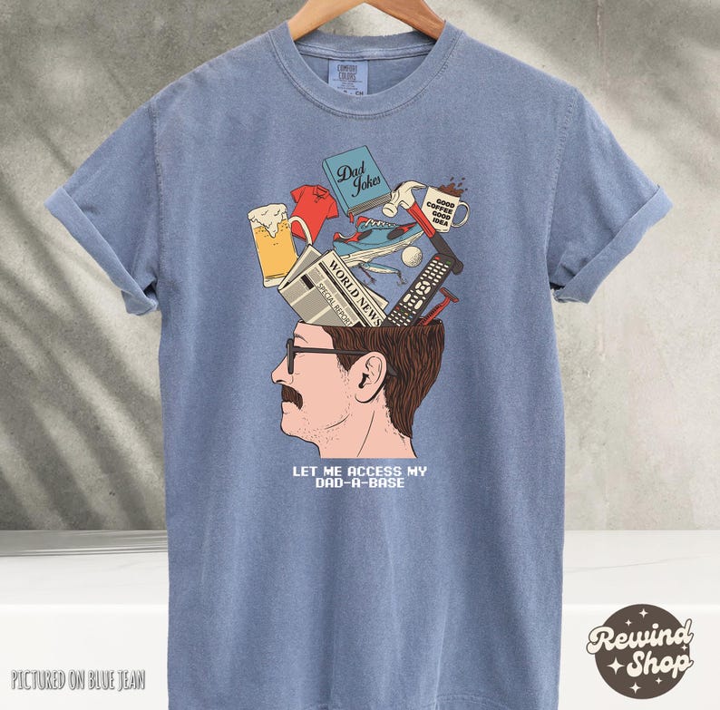 May include: Blue t-shirt with a graphic of a man's head, open to reveal various items like a beer, a remote, and a mug. The text "LET ME ACCESS MY DAD-A-BASE" is below the image. The shirt is displayed on a hanger.