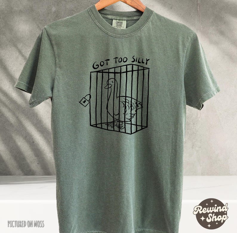 May include: A green t-shirt with a black and white illustration of a goose in a cage. The text "Got Too Silly" is printed above the image.