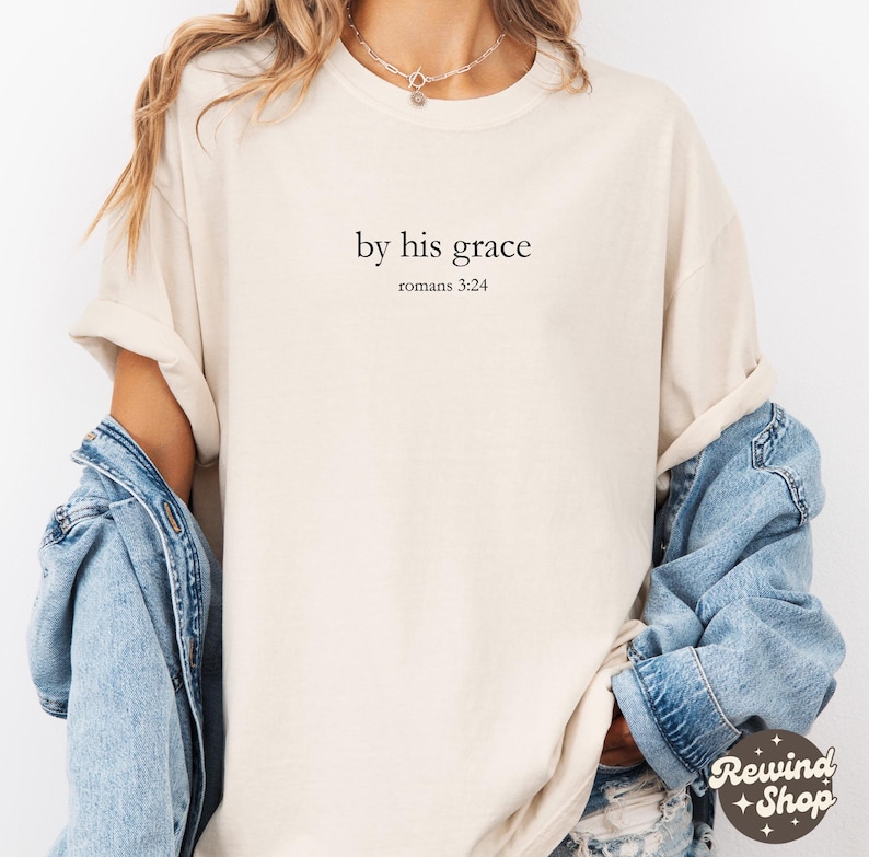 May include: A cream-coloured t-shirt featuring the text "by his grace" and "romans 3:24" in black. The shirt has short sleeves and a loose fit. A denim jacket is partially visible.