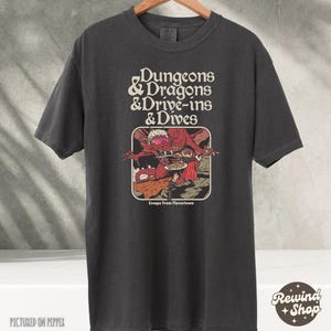 May include: Dark gray t-shirt featuring the text "Dungeons & Dragons & Drive-ins & Dives" above a graphic of a red dragon serving food. The graphic includes the text "Escape From Flavortown".