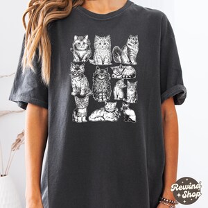 K&ouml;nnte beinhalten: Dunkelgraues T-Shirt mit einer Reihe detaillierter Schwarz-Wei&szlig;-Illustrationen von Katzen. Die Katzen sind in verschiedenen Posen dargestellt, darunter sitzen, liegen und stehen. Das Shirt hat eine lockere Passform und kurze &Auml;rmel.
