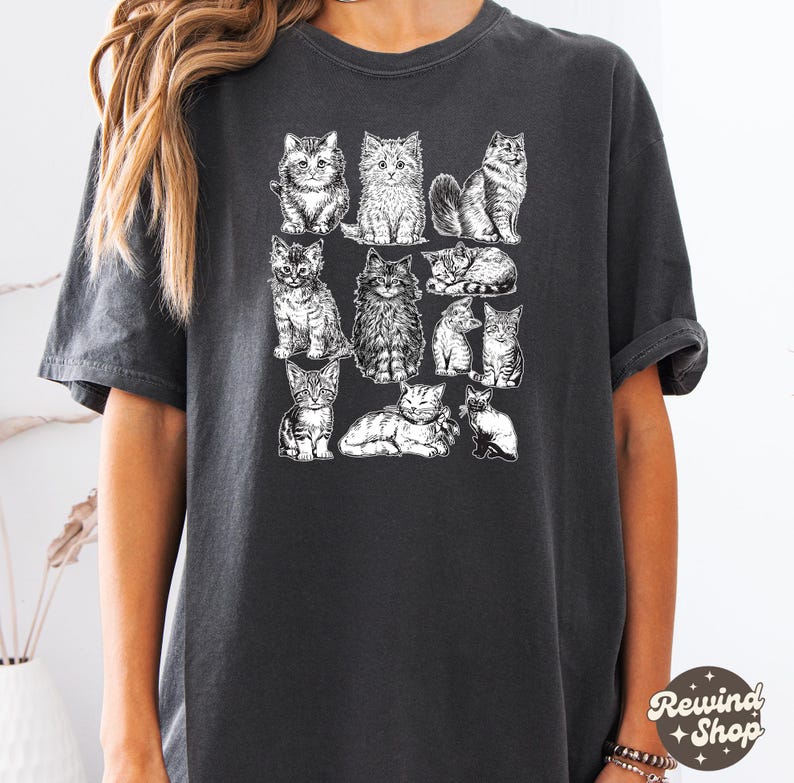 K&ouml;nnte beinhalten: Dunkelgraues T-Shirt mit einer Reihe detaillierter Schwarz-Wei&szlig;-Illustrationen von Katzen. Die Katzen sind in verschiedenen Posen dargestellt, darunter sitzen, liegen und stehen. Das Shirt hat eine lockere Passform und kurze &Auml;rmel.