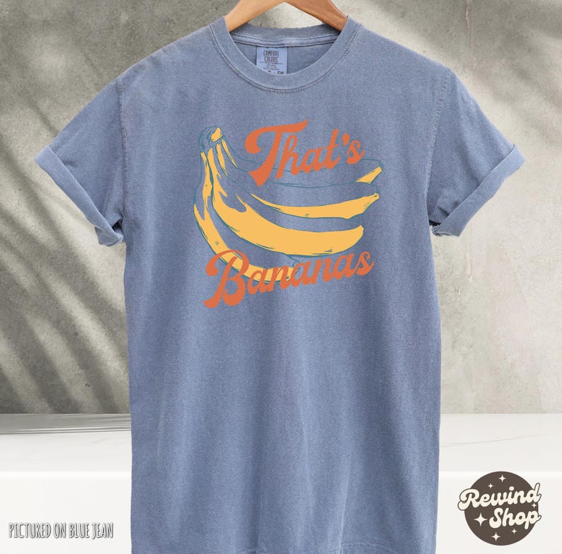 May include: Blue t-shirt with rolled sleeves featuring a yellow banana graphic and the text "That's Bananas" in orange. The shirt is displayed on a wooden hanger.