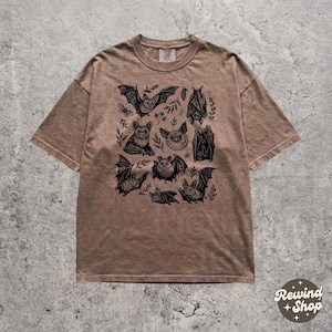May include: Brown t-shirt featuring a black illustration of various bats and foliage. The design includes bats in different poses, with spread wings and hanging upside down. The shirt has a relaxed fit and a crew neck.