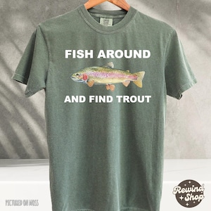 Pode incluir: T-shirt verde-oliva com o texto "FISH AROUND AND FIND TROUT" e uma ilustração de truta colorida. A t-shirt é exibida em um cabide de madeira.