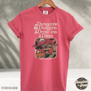 Może przedstawiać: Karmazynowa koszulka z podwiniętymi rękawami. Na koszulce widnieje napis "Dungeons & Dragons & Drive-ins & Dives" oraz grafika smoka i osoby. Pod grafiką znajduje się napis "Escape From Flavortown". Koszulka wisi na drewnianym wieszaku.