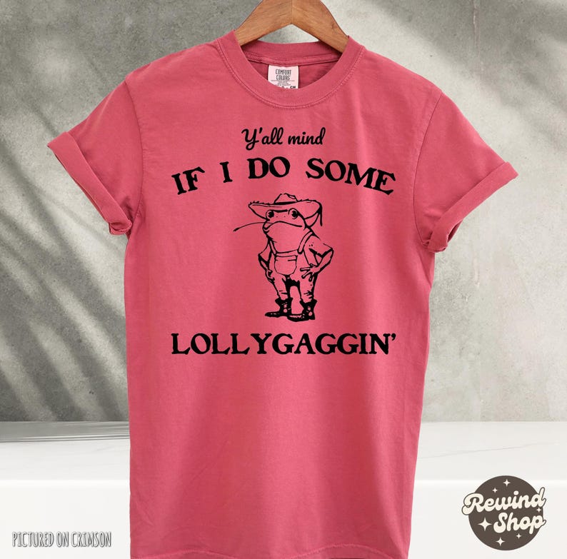 Op de afbeelding: Een roze T-shirt met een zwarte afbeelding van een kikker die een hoed en een overall draagt. De tekst op het shirt luidt "Y'all mind IF I DO SOME LOLLYGAGGIN'"