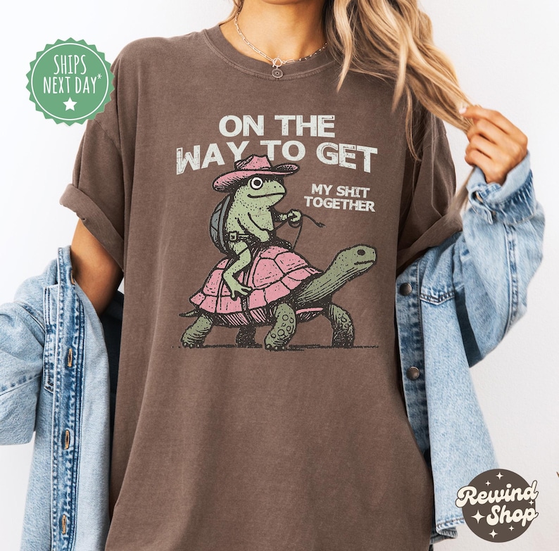 May include: Brown t-shirt with a graphic of a frog riding a turtle. The text on the shirt reads "On the way to get my shit together."