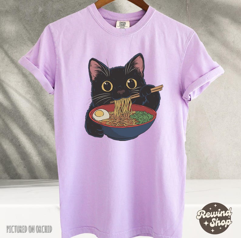 May include: A purple t-shirt with a black cat eating ramen noodles. The cat is wearing a red collar and has a bowl of ramen in front of it. The text "Rewind Shop" is in the bottom right corner of the image.
