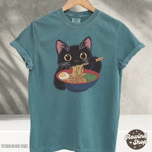 May include: A teal blue t-shirt with a black cat eating ramen noodles. The cat is wearing a red collar and has a white egg in its bowl.