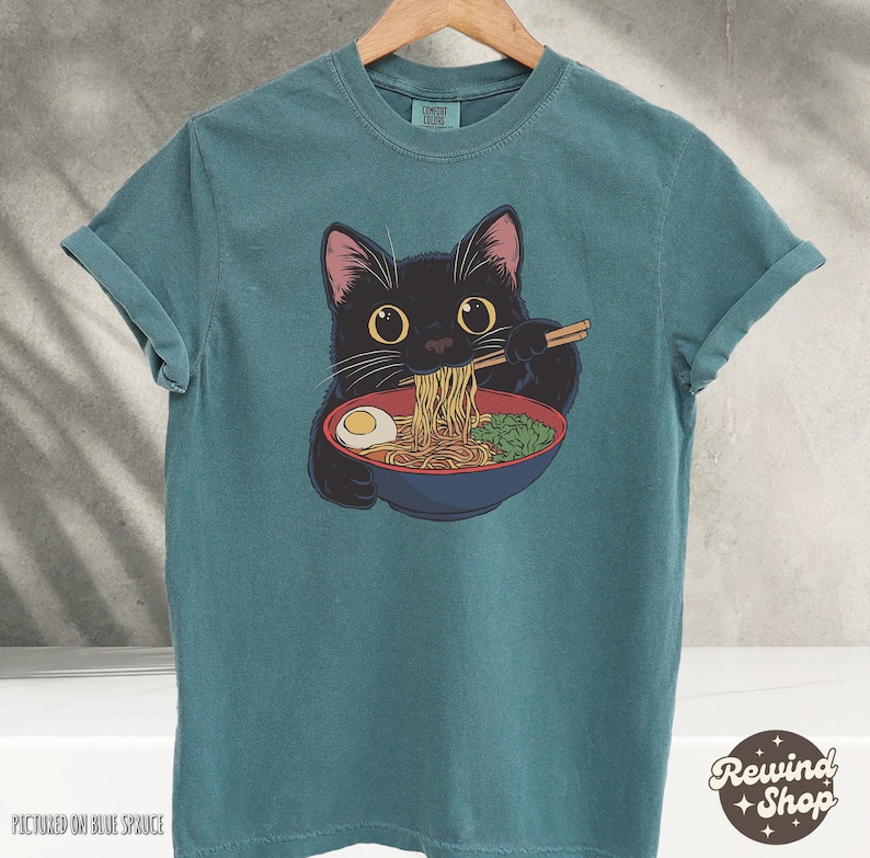 May include: A teal blue t-shirt with a black cat eating ramen noodles. The cat is wearing a red collar and has a white egg in its bowl.