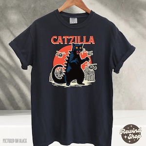 May include: Black t-shirt with a graphic of a black cat monster with red eyes, a city skyline, and the text "CATZILLA".