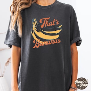 May include: Dark gray t-shirt featuring a graphic of yellow bananas and the text "That's Bananas" in orange. The shirt has a vintage, washed look and a relaxed fit, suitable for casual wear.