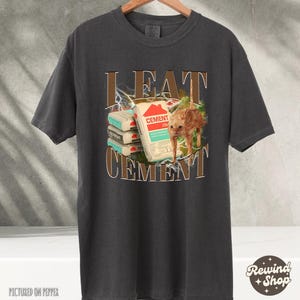 May include: A dark gray t-shirt with the text "I EAT CEMENT" in brown letters. The text is surrounded by a lightning bolt design. A cat is sitting on top of a stack of cement bags.