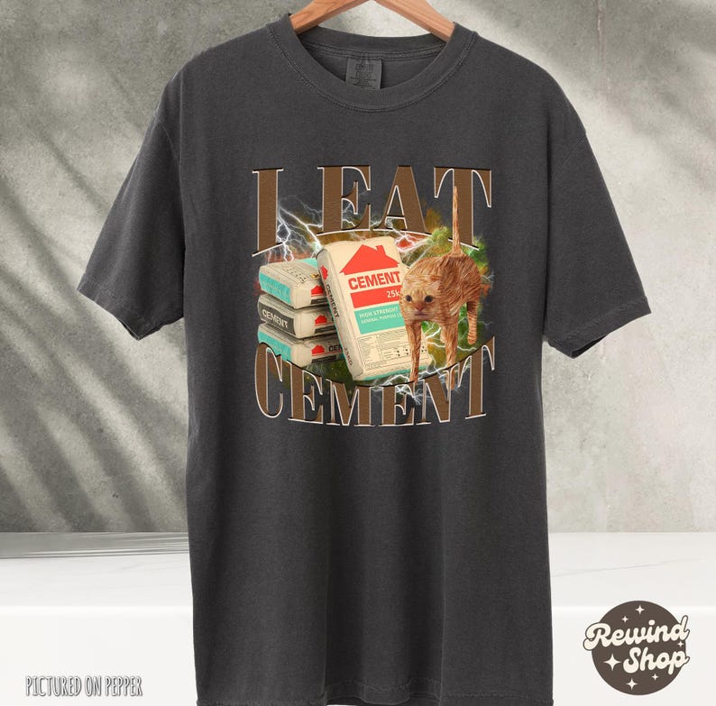 May include: A dark gray t-shirt with the text "I EAT CEMENT" in brown letters. The text is surrounded by a lightning bolt design. A cat is sitting on top of a stack of cement bags.