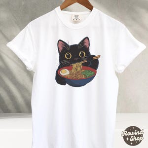May include: White t-shirt with a black cat eating ramen noodles. The cat is wearing a red bowl and holding chopsticks.