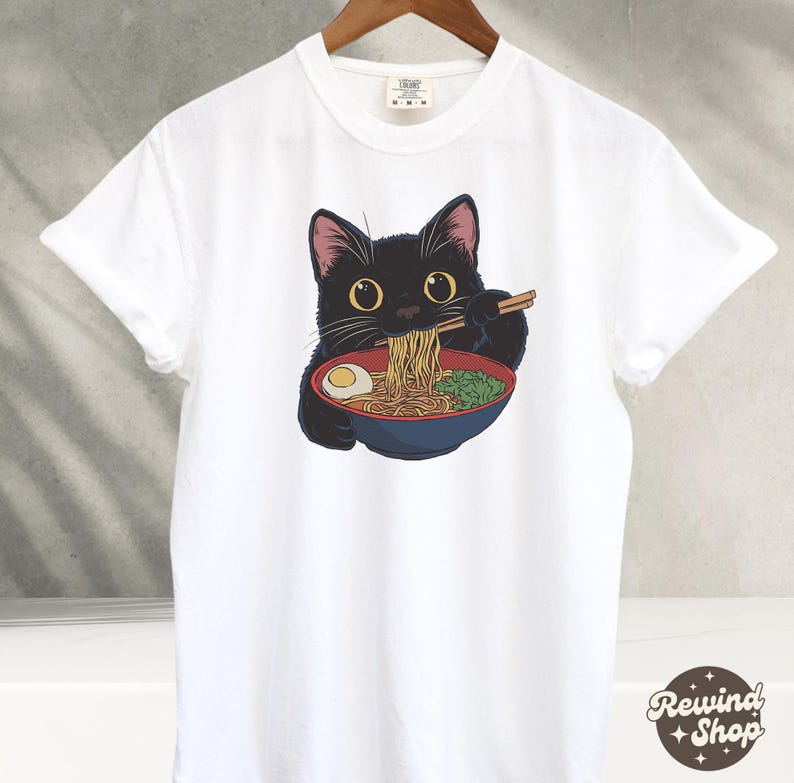 May include: White t-shirt with a black cat eating ramen noodles. The cat is wearing a red bowl and holding chopsticks.
