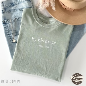 May include: A sage green t-shirt with the text "by his grace romans 3:24" in white. The shirt is laid on top of light blue jeans, with a gold necklace and a tan hat. The image is from Rewind Shop.